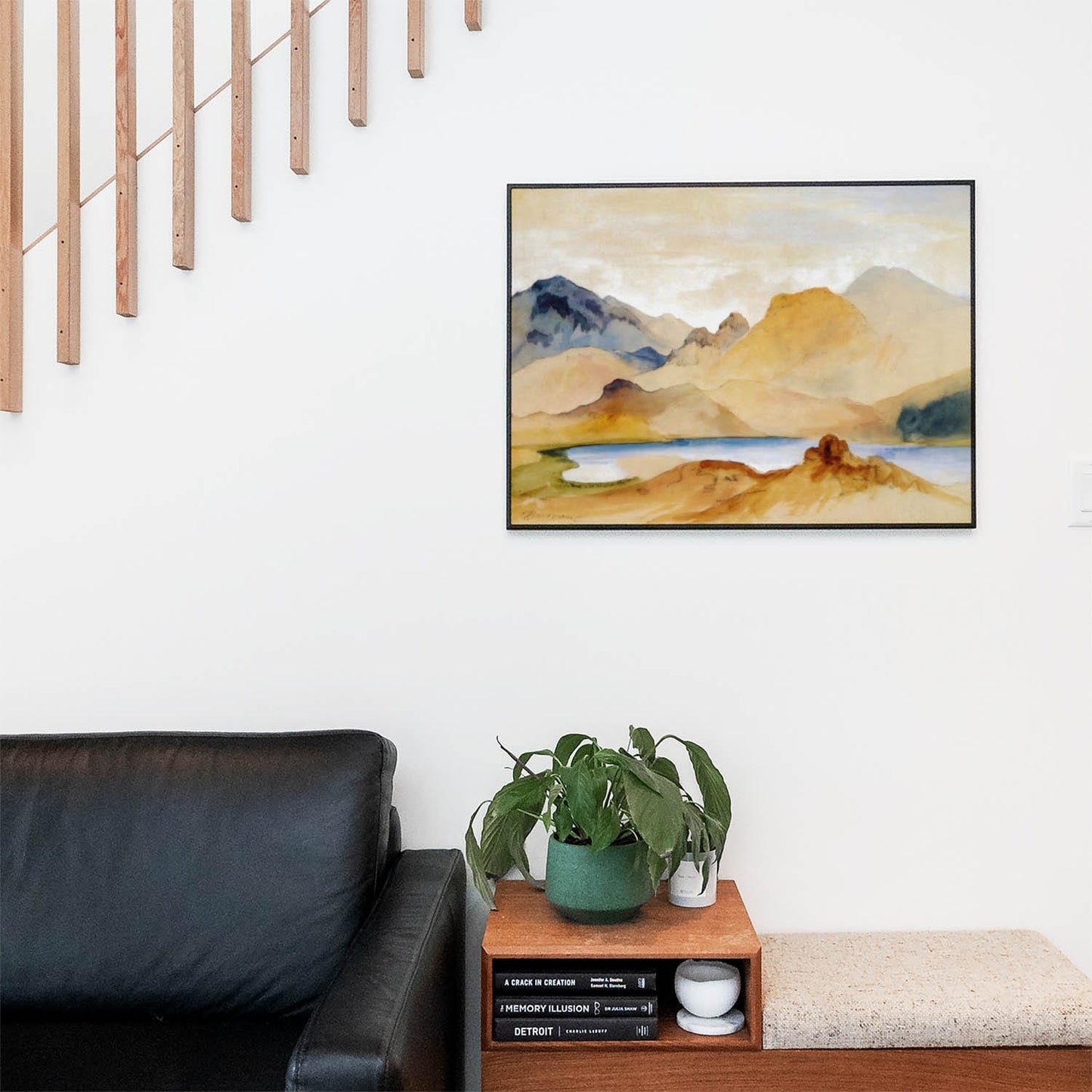 Living space with a black leather couch and table with a plant and books below a staircase featuring a framed picture of Nature Landscape