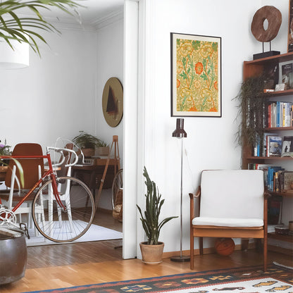 Eclectic living room with a road bike, bookshelf and house plants that features framed artwork of a Bohemain Floral above a chair and lamp