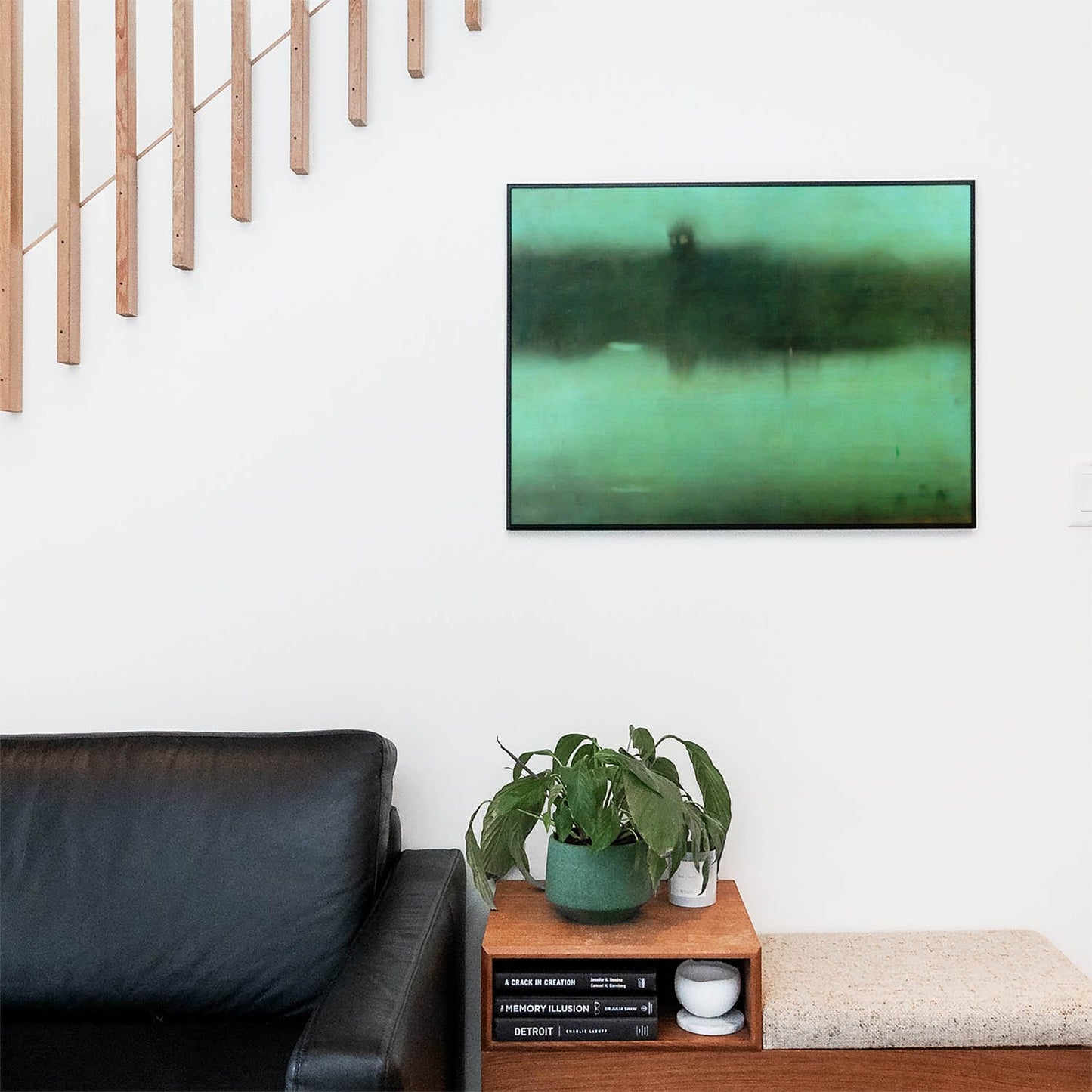 Living space with a black leather couch and table with a plant and books below a staircase featuring a framed picture of Moody Abstract