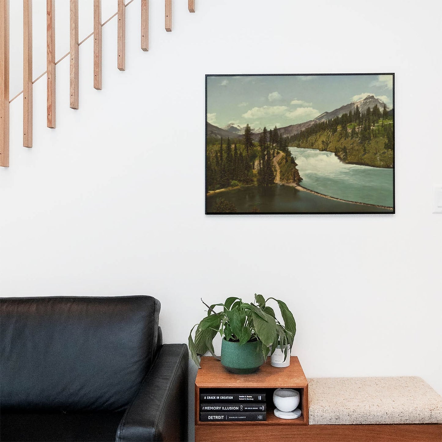 Living space with a black leather couch and table with a plant and books below a staircase featuring a framed picture of Mountains and Rivers