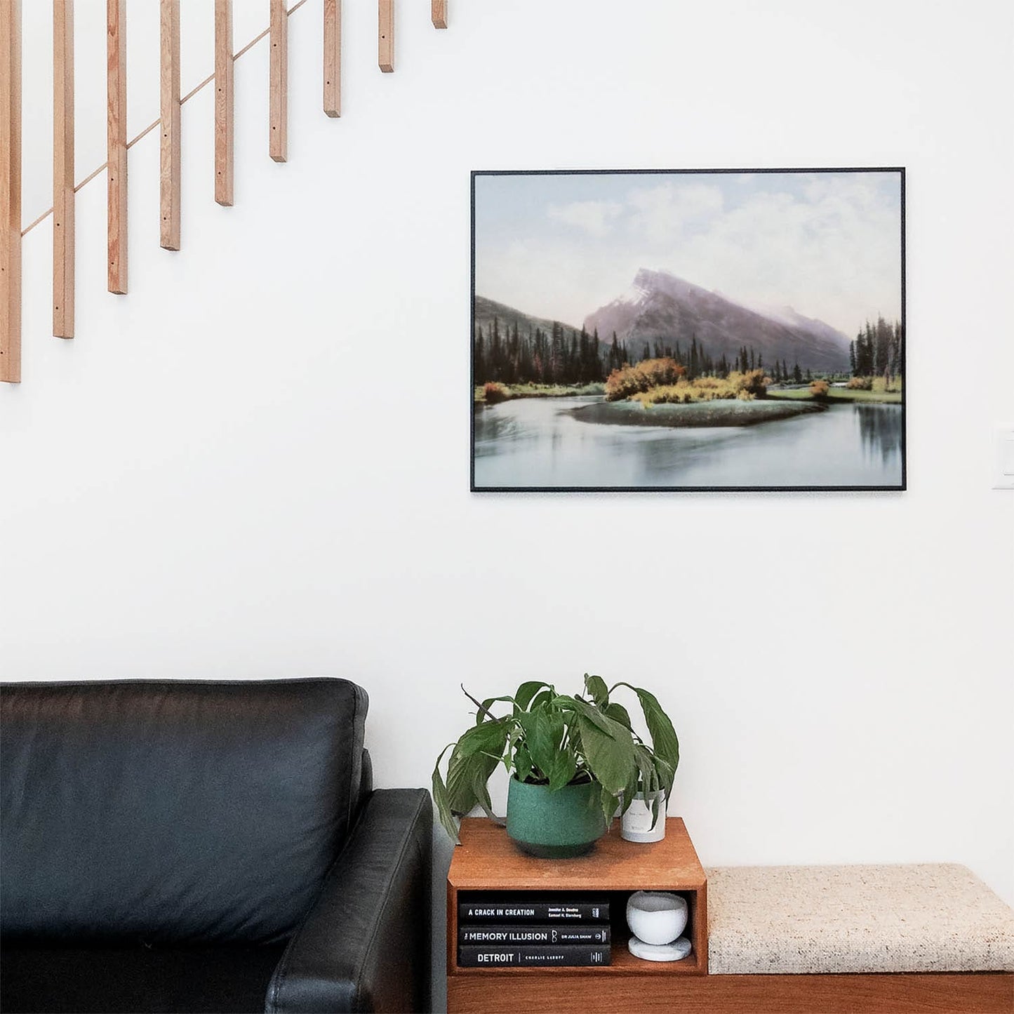 Living space with a black leather couch and table with a plant and books below a staircase featuring a framed picture of Mountain and Lake