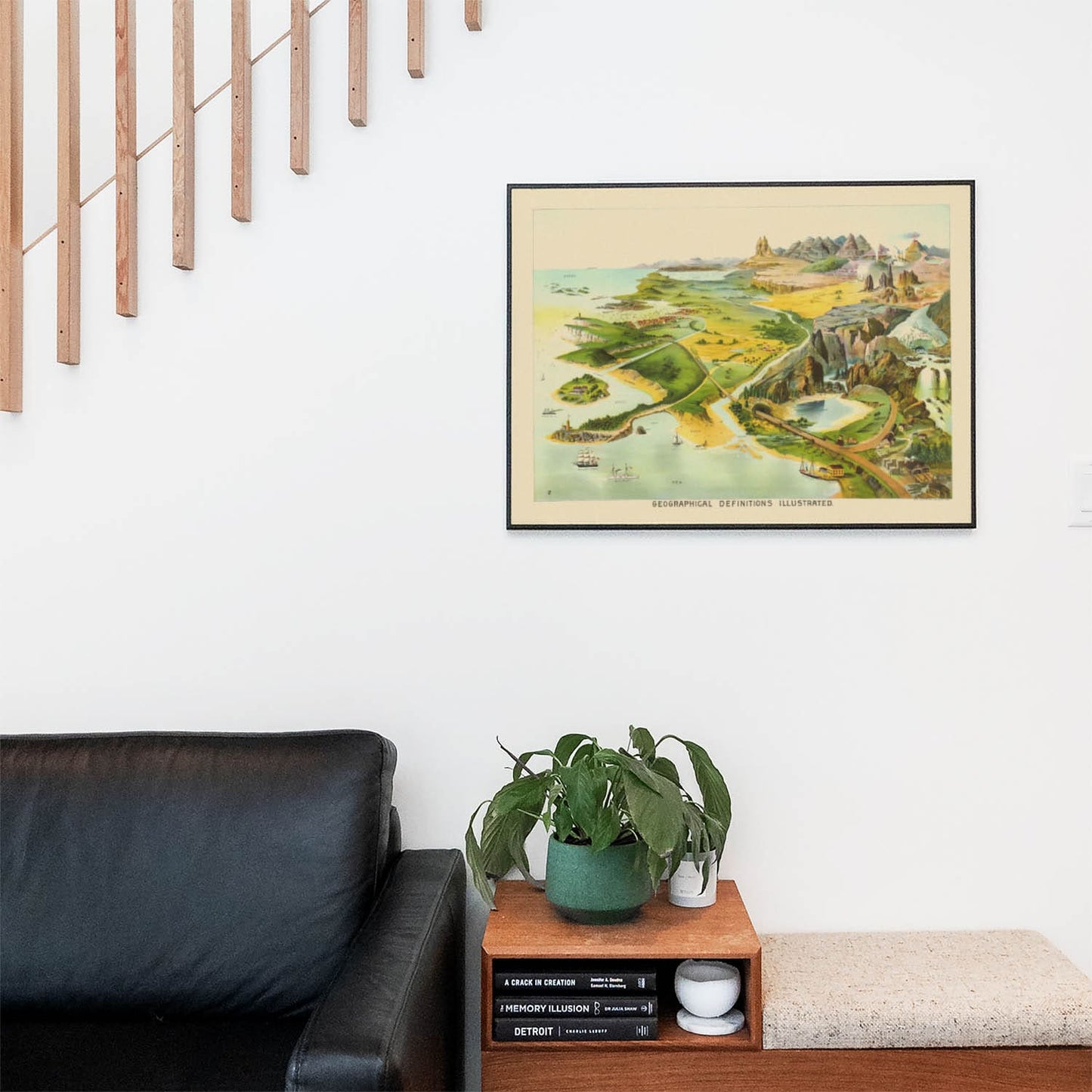 Living space with a black leather couch and table with a plant and books below a staircase featuring a framed picture of Vintage Geography