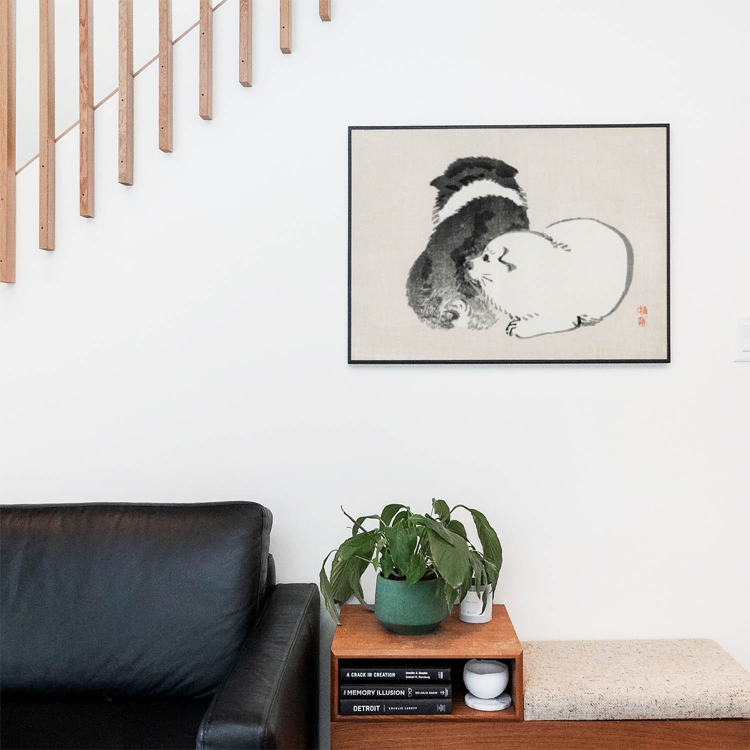 Living space with a black leather couch and table with a plant and books below a staircase featuring a framed picture of Minimalist Puppy