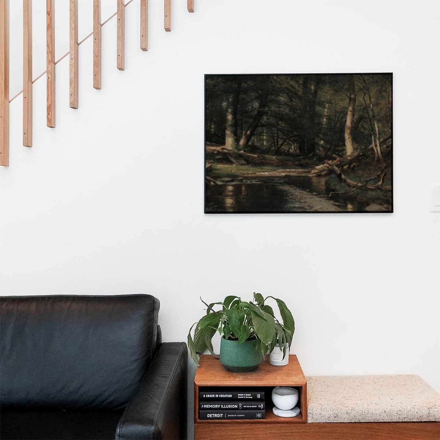 Living space with a black leather couch and table with a plant and books below a staircase featuring a framed picture of Dark Nature