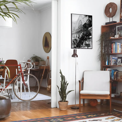 Eclectic living room with a road bike, bookshelf and house plants that features framed artwork of a Train Falling from a Building above a chair and lamp