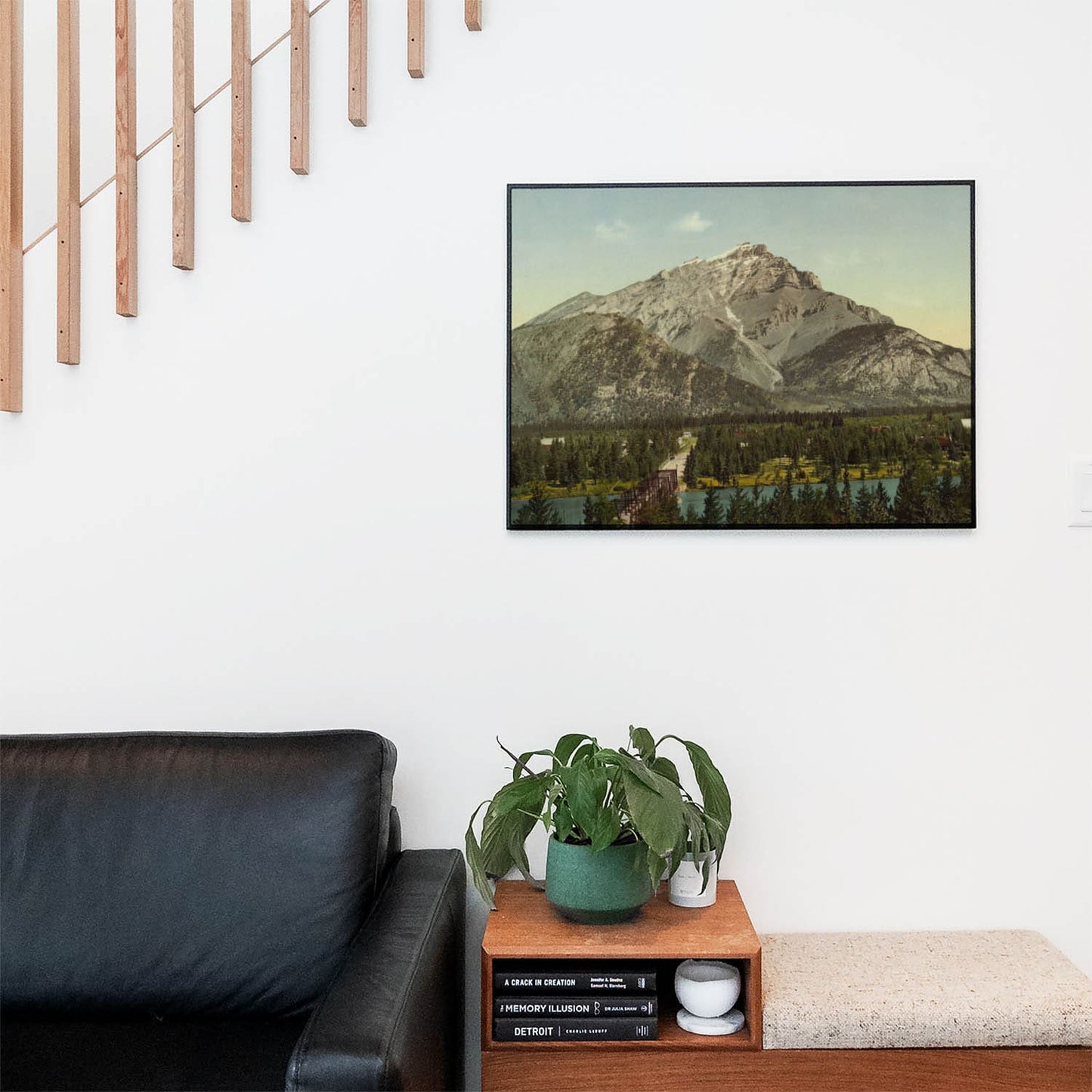 Living space with a black leather couch and table with a plant and books below a staircase featuring a framed picture of Vintage Mountains
