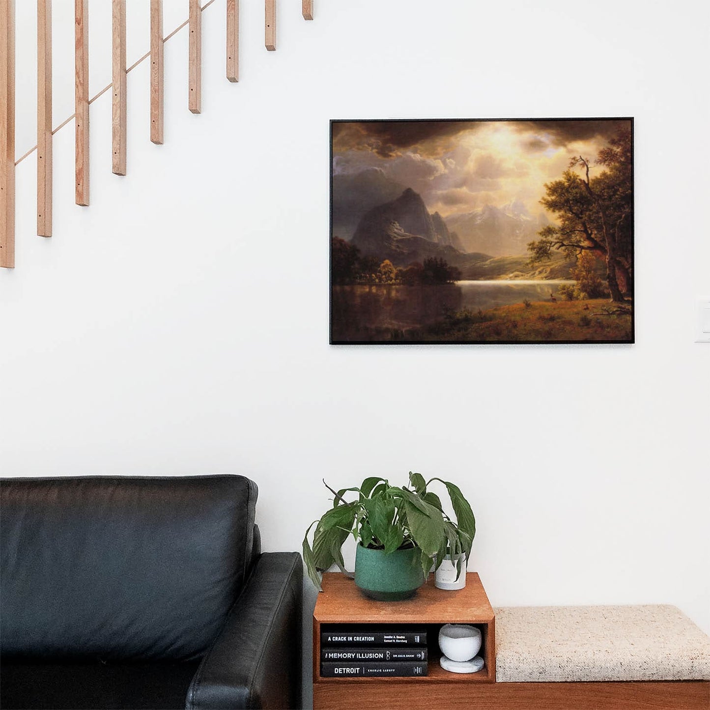 Living space with a black leather couch and table with a plant and books below a staircase featuring a framed picture of Mountian and Lake