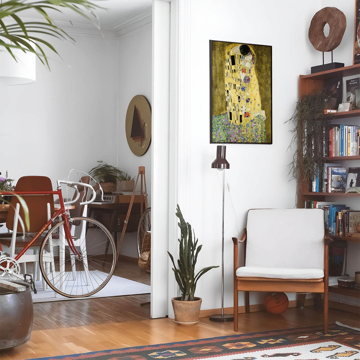 Eclectic living room with a road bike, bookshelf and house plants that features framed artwork of a Two Lovers Embracing in a Kiss Wearing Gold Robes above a chair and lamp