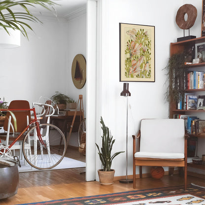 Eclectic living room with a road bike, bookshelf and house plants that features framed artwork of a Aesthetic Watercolor above a chair and lamp