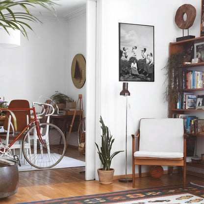 Eclectic living room with a road bike, bookshelf and house plants that features framed artwork of a Vintage Classy Women Drinking Alcohol above a chair and lamp