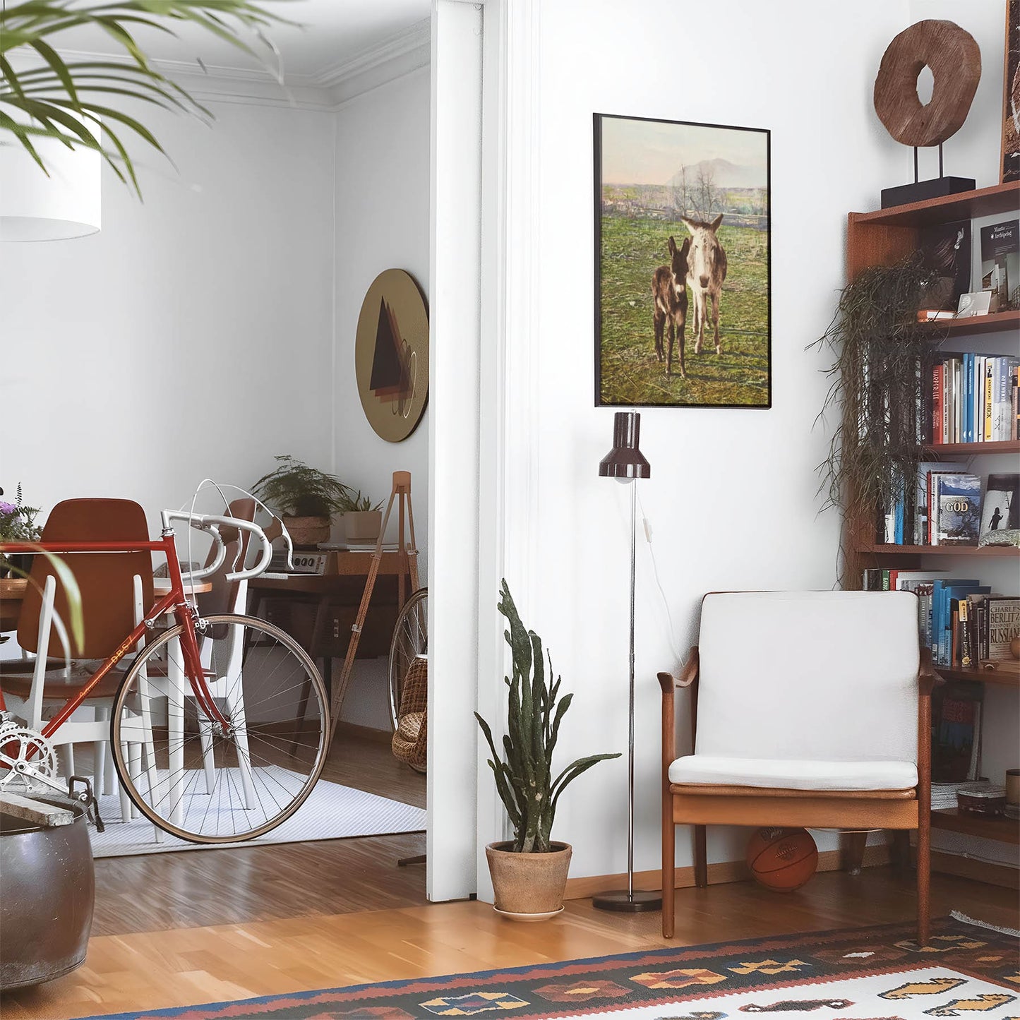 Eclectic living room with a road bike, bookshelf and house plants that features framed artwork of a Two Donkey's in the Mountains above a chair and lamp