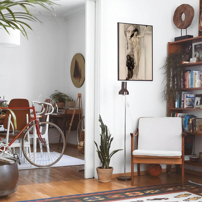 Eclectic living room with a road bike, bookshelf and house plants that features framed artwork of a Female Figure Sculpture above a chair and lamp