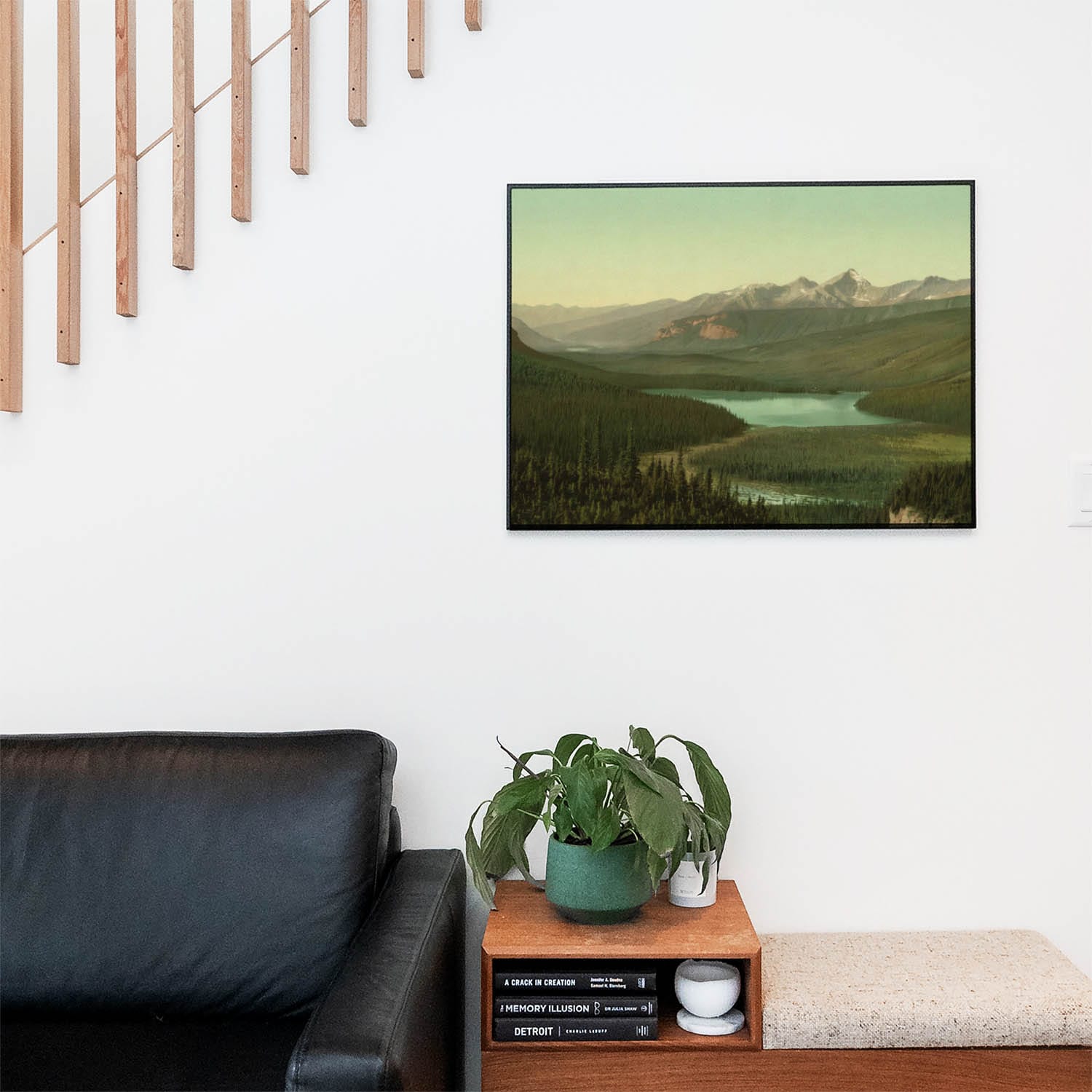 Living space with a black leather couch and table with a plant and books below a staircase featuring a framed picture of Emerald Green Landscape