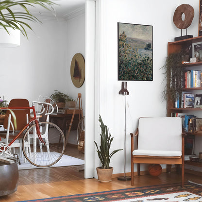 Eclectic living room with a road bike, bookshelf and house plants that features framed artwork of a Green, Pink and Red Flowers above a chair and lamp
