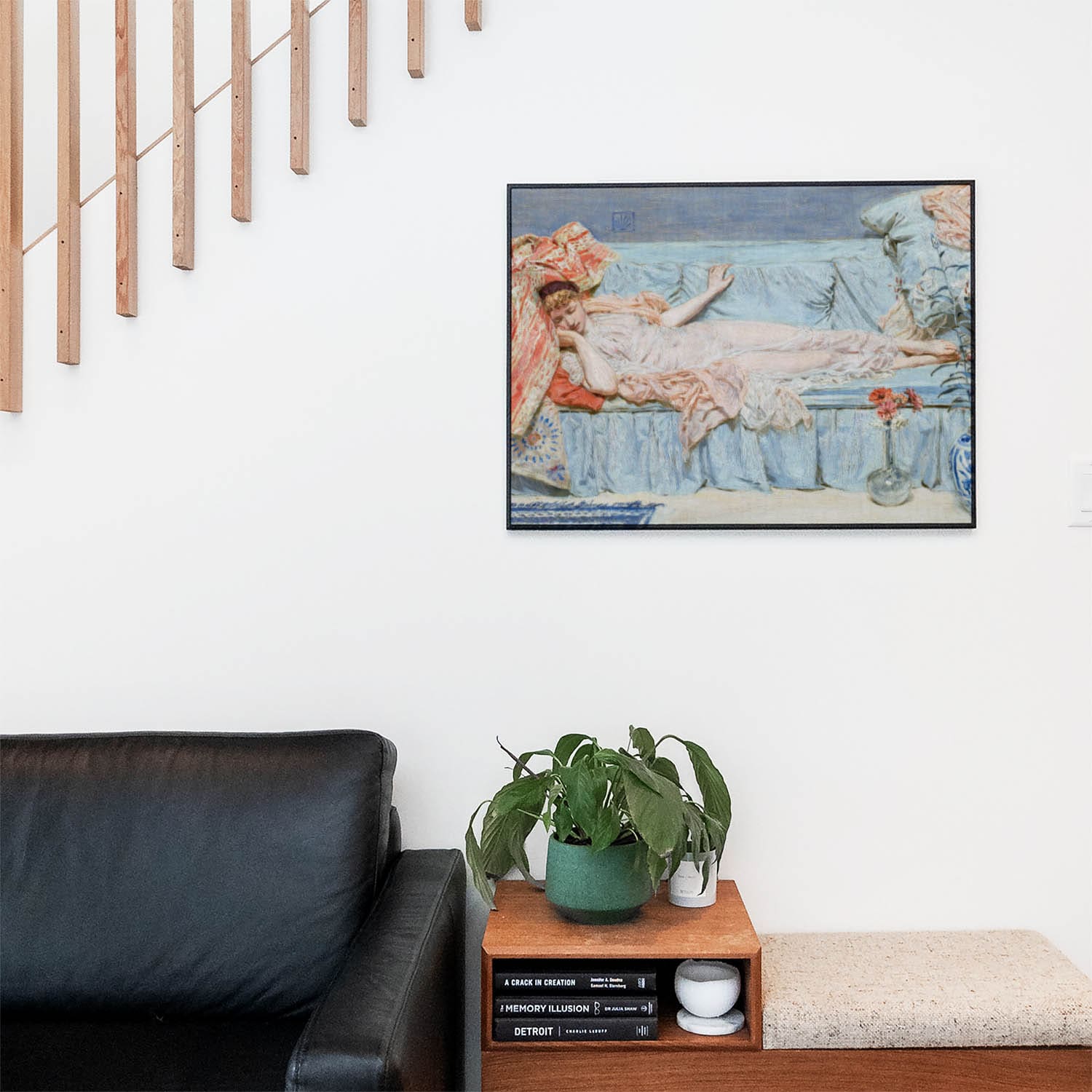 Living space with a black leather couch and table with a plant and books below a staircase featuring a framed picture of Asleep on a Blue Couch