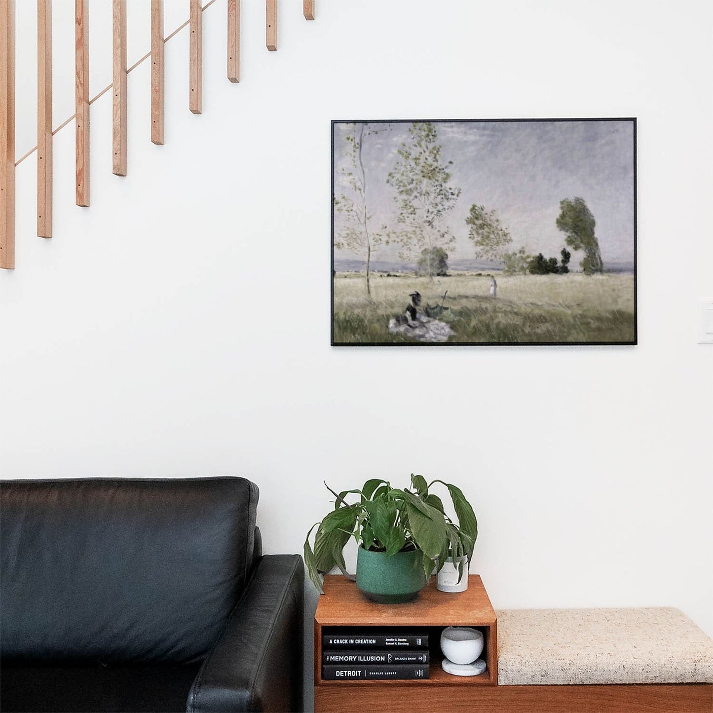 Living space with a black leather couch and table with a plant and books below a staircase featuring a framed picture of Seeking Time Alone