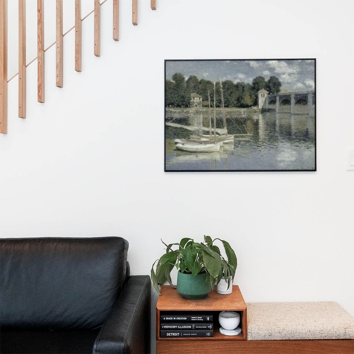 Living space with a black leather couch and table with a plant and books below a staircase featuring a framed picture of Sage Green Bridge