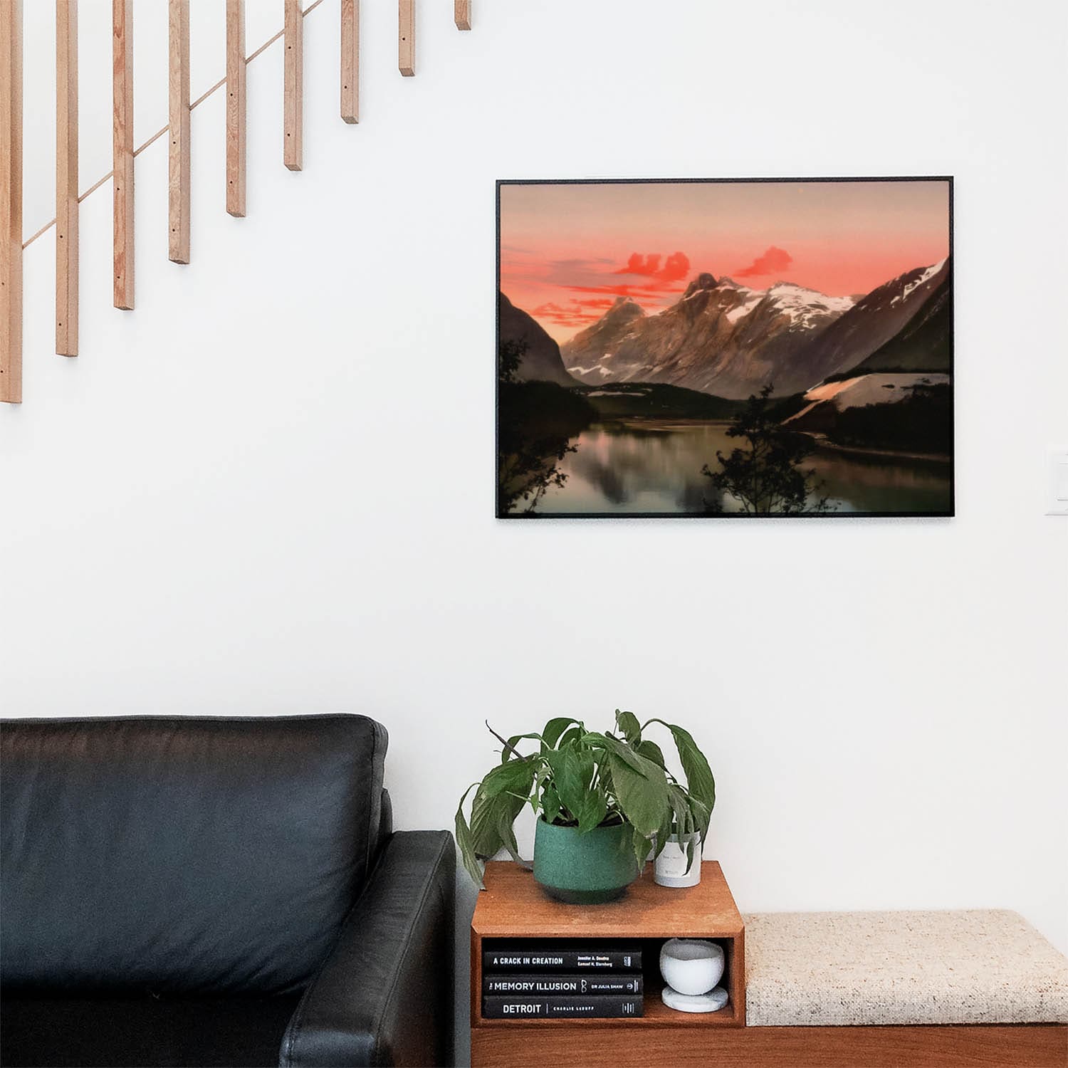 Living space with a black leather couch and table with a plant and books below a staircase featuring a framed picture of Mountains and Lake Nature
