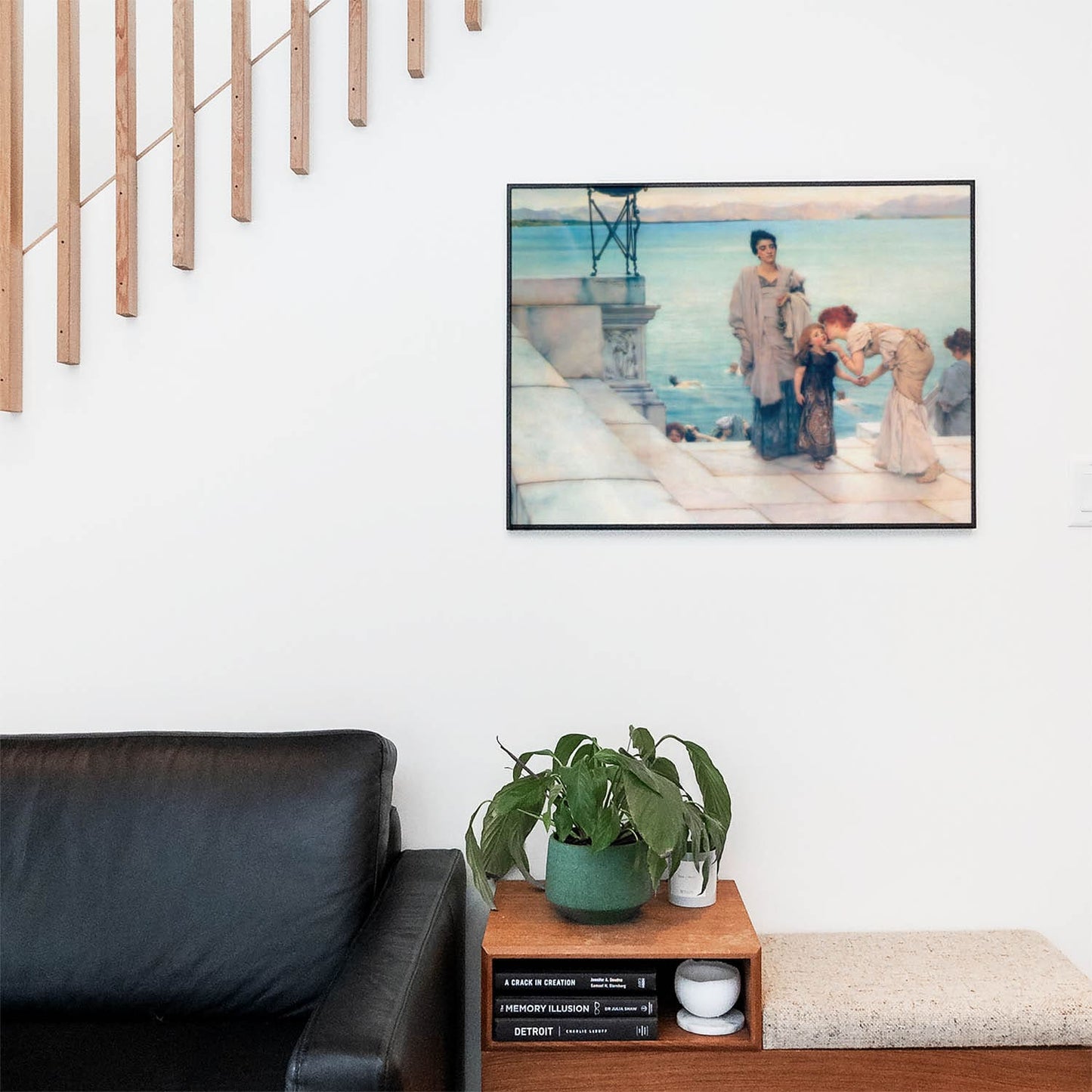 Living space with a black leather couch and table with a plant and books below a staircase featuring a framed picture of At the Lake