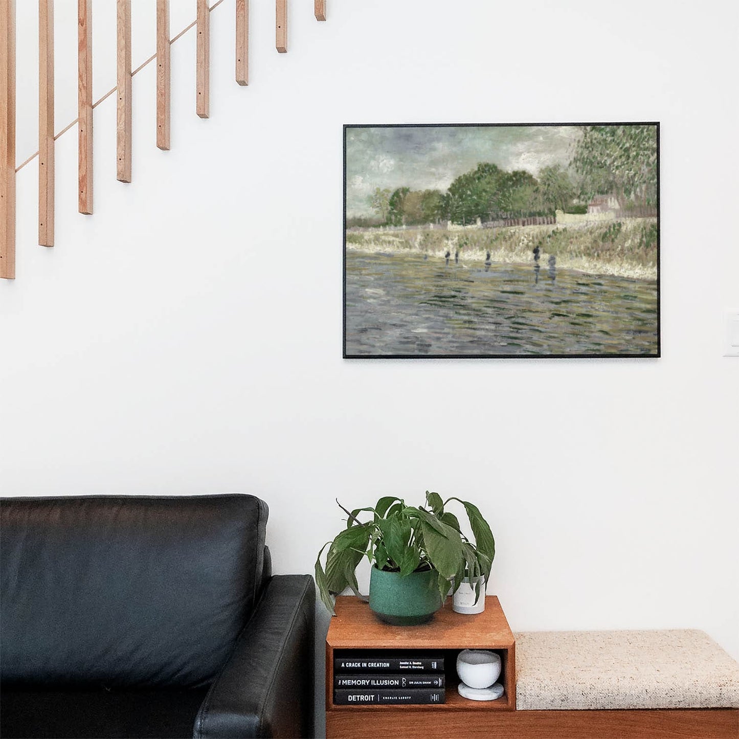 Living space with a black leather couch and table with a plant and books below a staircase featuring a framed picture of Lush Greenery