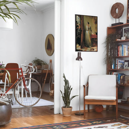 Eclectic living room with a road bike, bookshelf and house plants that features framed artwork of a Victorian Era Mystery above a chair and lamp