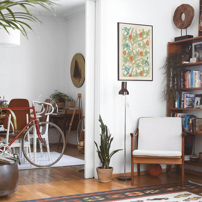 Eclectic living room with a road bike, bookshelf and house plants that features framed artwork of a Plants and Birds above a chair and lamp