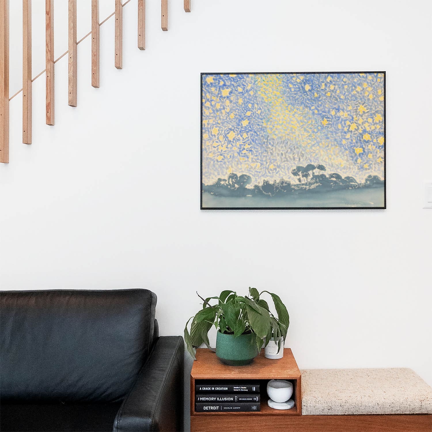 Living space with a black leather couch and table with a plant and books below a staircase featuring a framed picture of Blue and Yellow