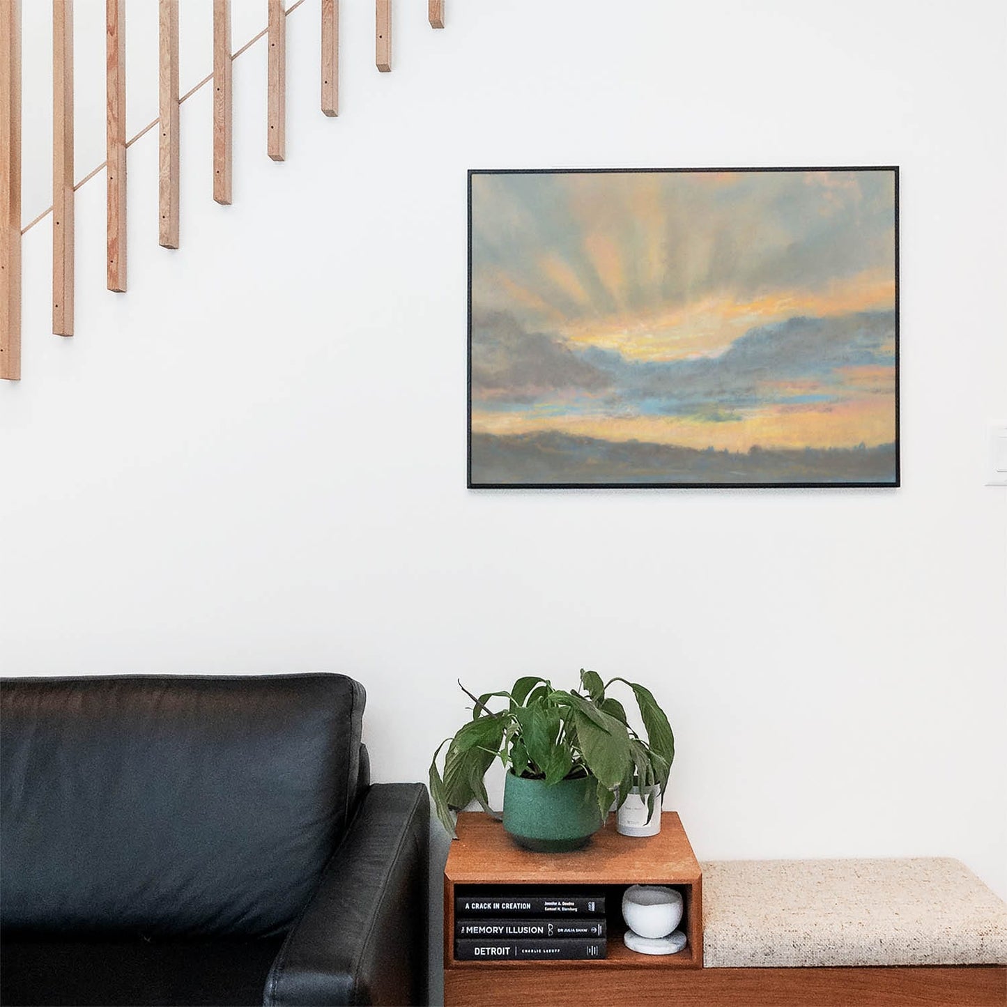 Living space with a black leather couch and table with a plant and books below a staircase featuring a framed picture of Yellow and Blue Sunset