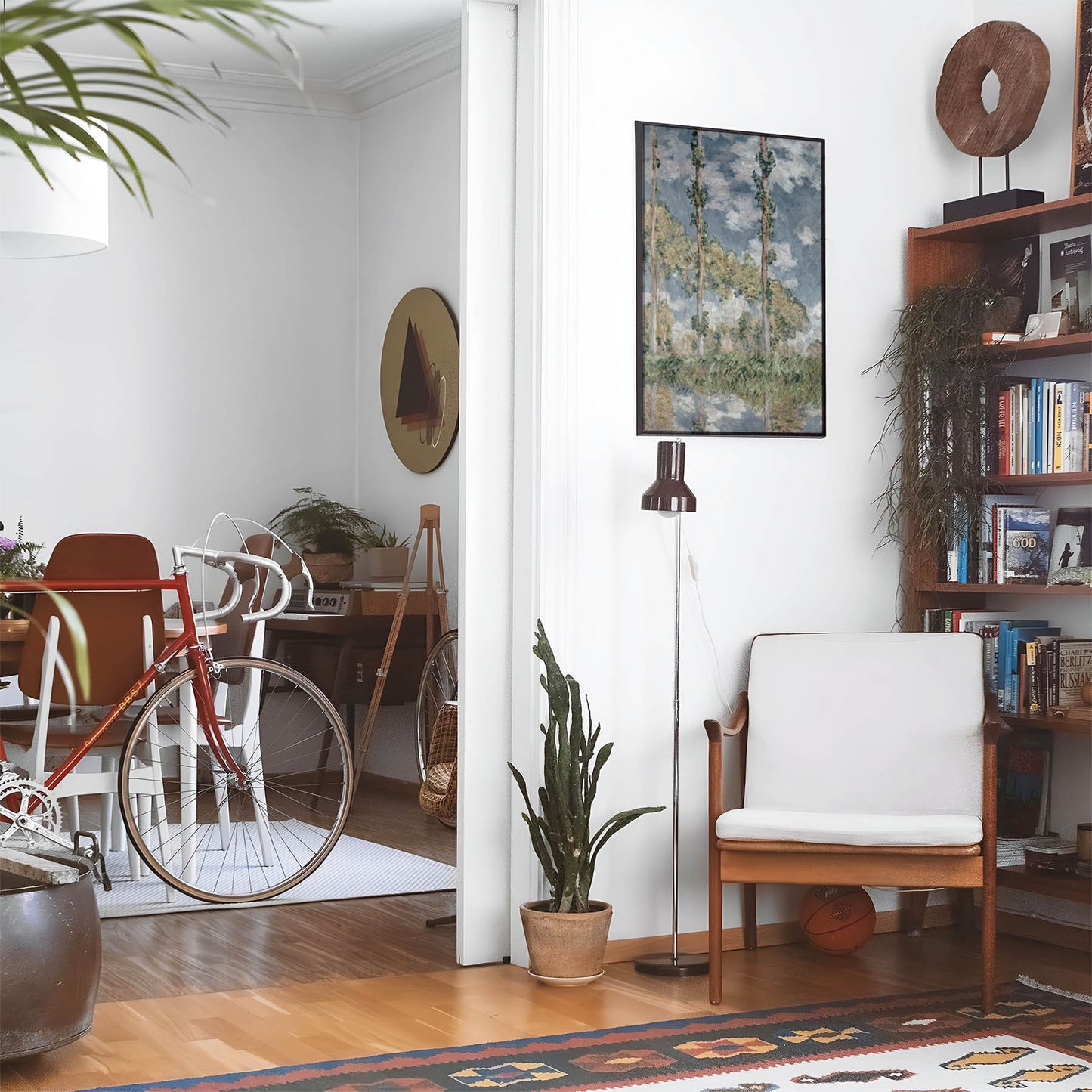 Eclectic living room with a road bike, bookshelf and house plants that features framed artwork of a Trees and Sky above a chair and lamp