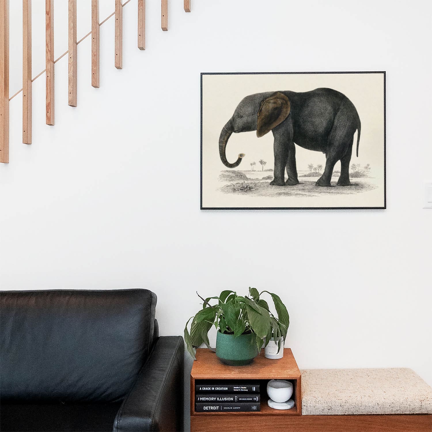 Living space with a black leather couch and table with a plant and books below a staircase featuring a framed picture of Cute Elephant
