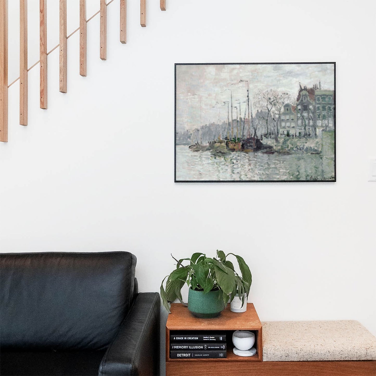 Living space with a black leather couch and table with a plant and books below a staircase featuring a framed picture of Seascape and City