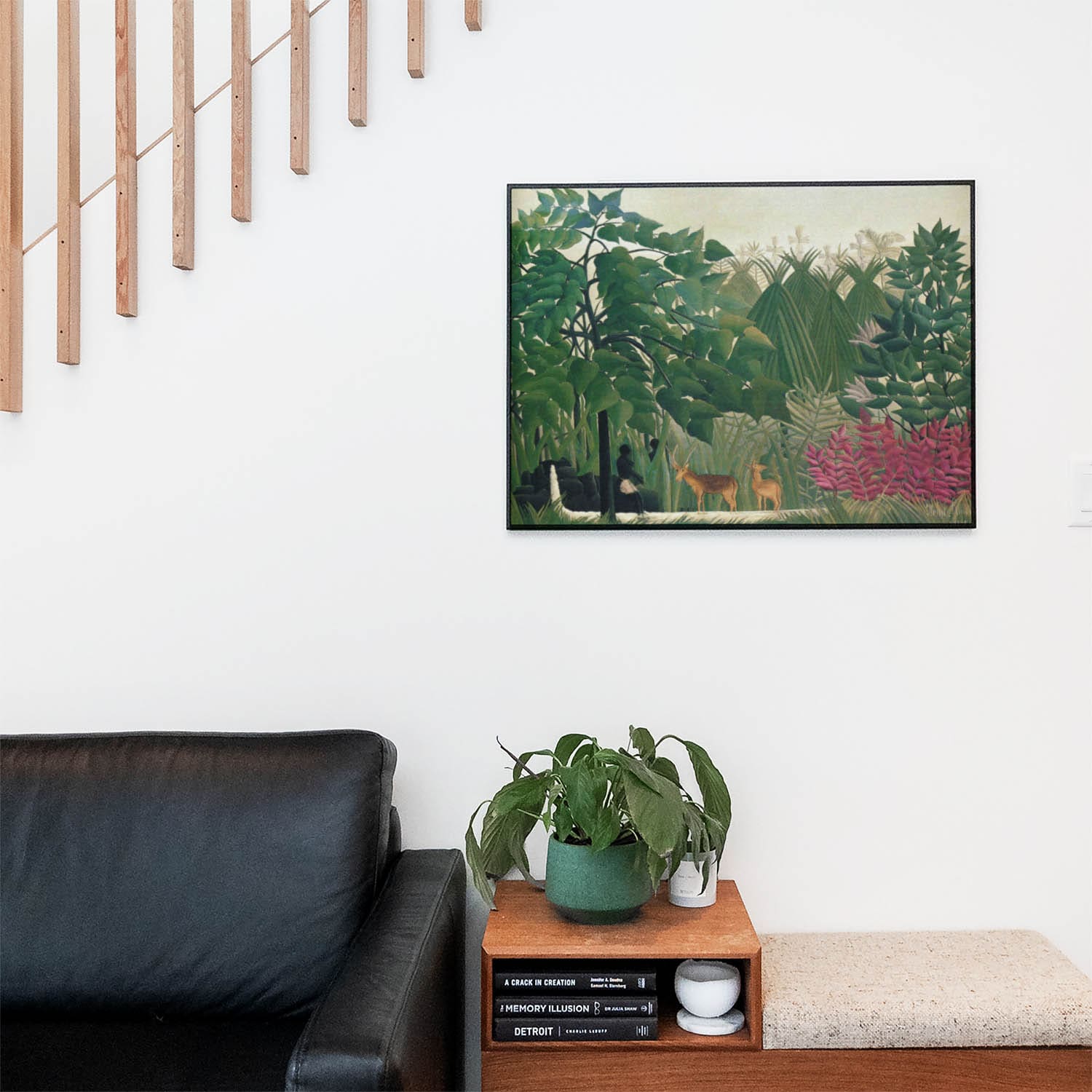 Living space with a black leather couch and table with a plant and books below a staircase featuring a framed picture of Tropical